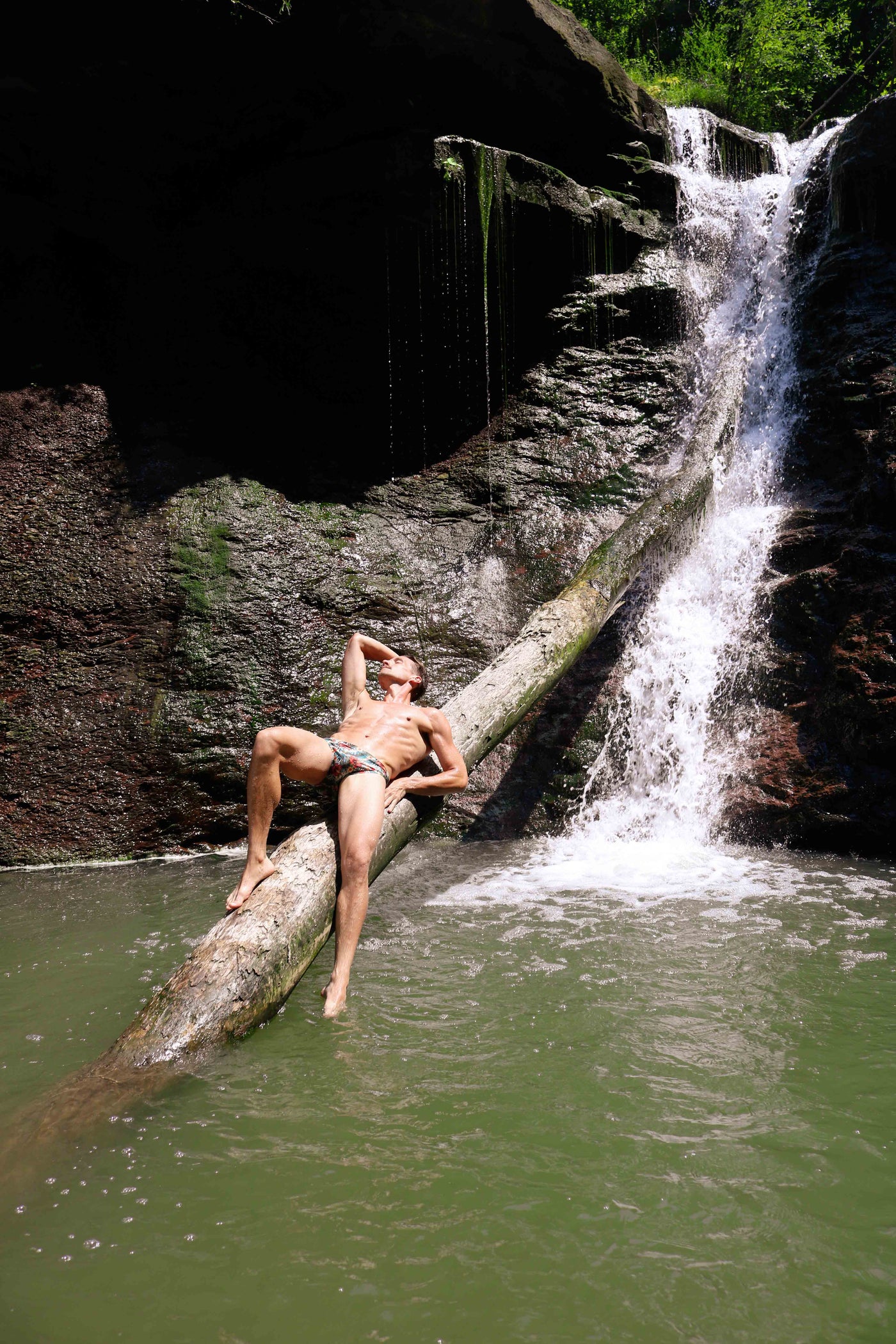 Man reclining on a log at the base of a waterfall, wearing HAC Swim's Hibiscus print swim brief.