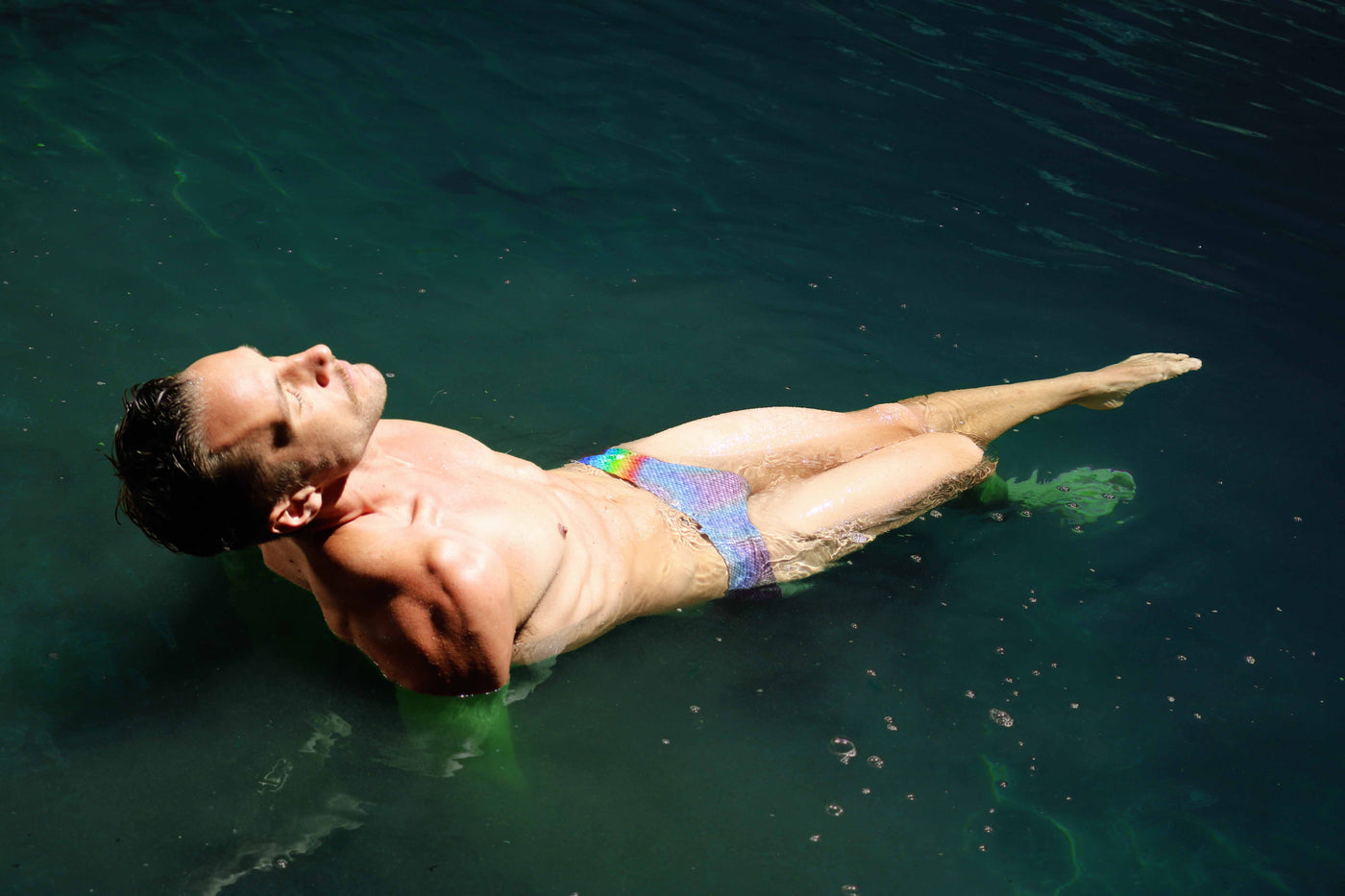 Man floating on his back in dark green water, wearing HAC Swim's Rainbow Trout swim brief, eyes closed.