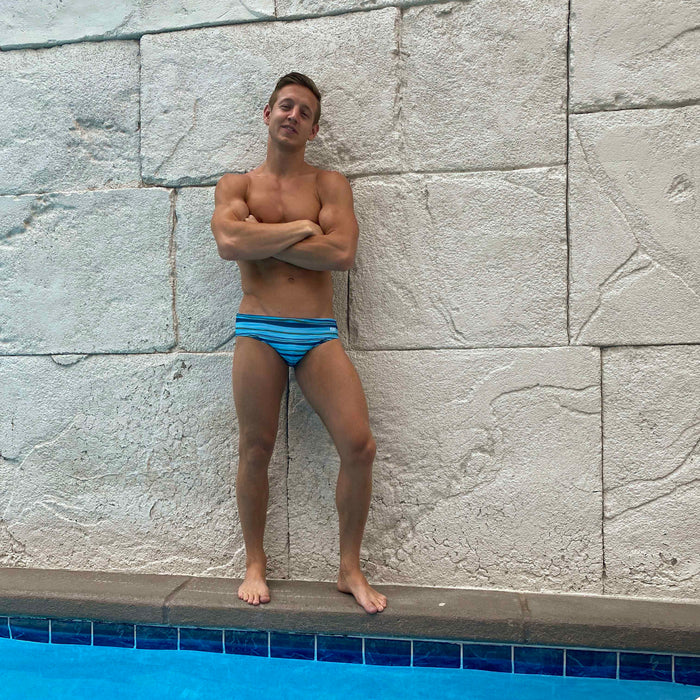 Model wearing the HAC Swim Tidal Stripe Basic swim brief beside a pool and textured stone wall.